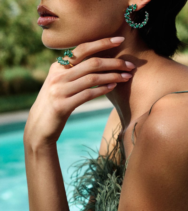 A close-up lifestyle shot of a model wearing a matching green gemstone ring and hoop earring, with a turquoise swimming pool and lush green background.