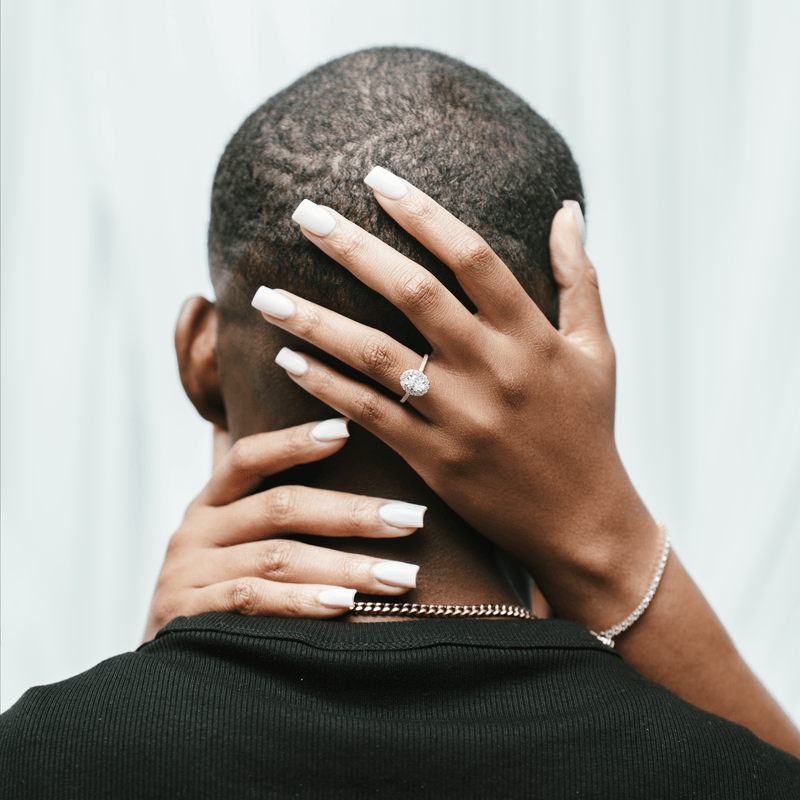 A close-up lifestyle shot from behind of a person with short dark hair, wearing a black shirt and a gold chain necklace, with another person's hands resting on their head. The person wearing the ring has long white manicured nails and a silver ring with a clear gemstone.
