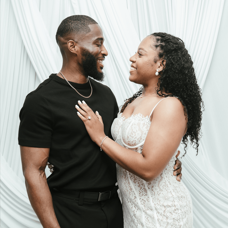 A couple posing together in front of a draped white fabric backdrop. The man is wearing a black t-shirt and black pants, and the woman is wearing a white lace spaghetti-strap dress.