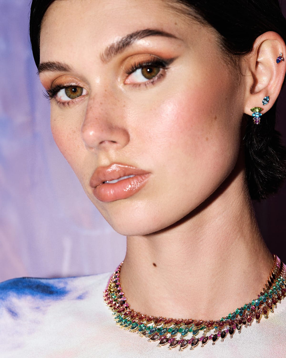 A close-up portrait of a model wearing a multi-strand statement necklace and matching drop earrings. The jewelry features small, colorful gemstones in shades of green, blue, pink, and gold, set against a soft, out-of-focus purple background.