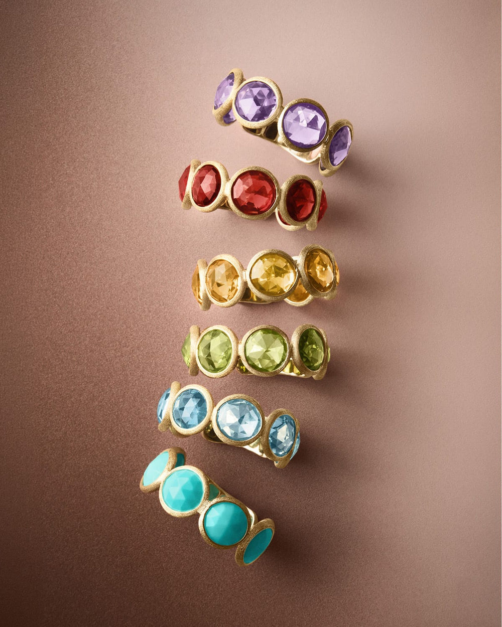 Six gold rings with round gemstones in purple, red, yellow, green, blue, and turquoise, arranged vertically on a warm brown background.
