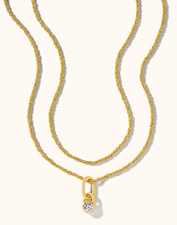 A gold double-layer necklace featuring a textured chain and a small pendant with a bezel-set clear gemstone, displayed on a light cream background.