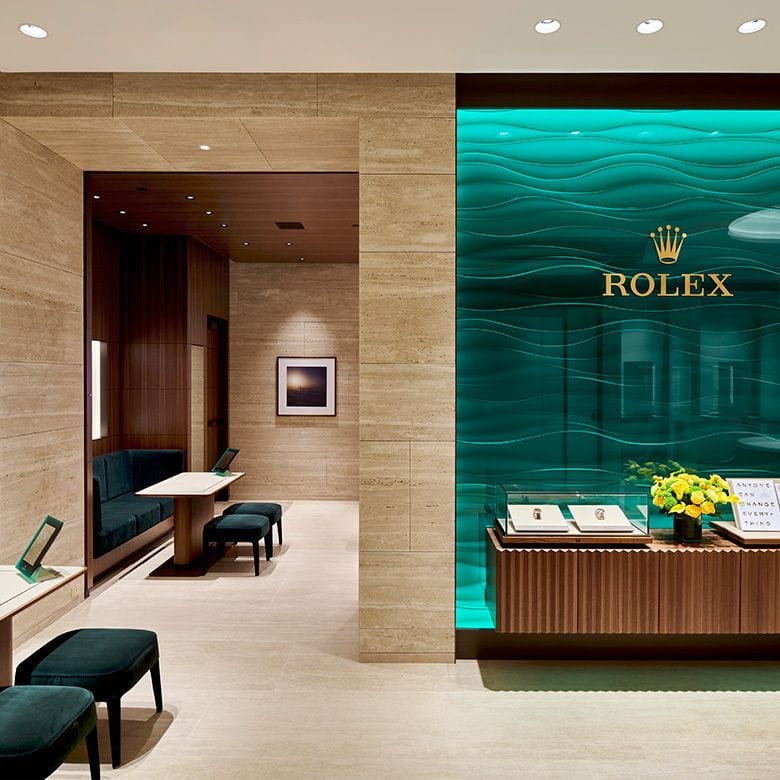 A luxurious Rolex store interior featuring a display case with watches, a teal accent wall with the Rolex logo, and elegant wood paneling. There are seating areas with green upholstered benches and small tables. Yellow flowers adorn the display case.