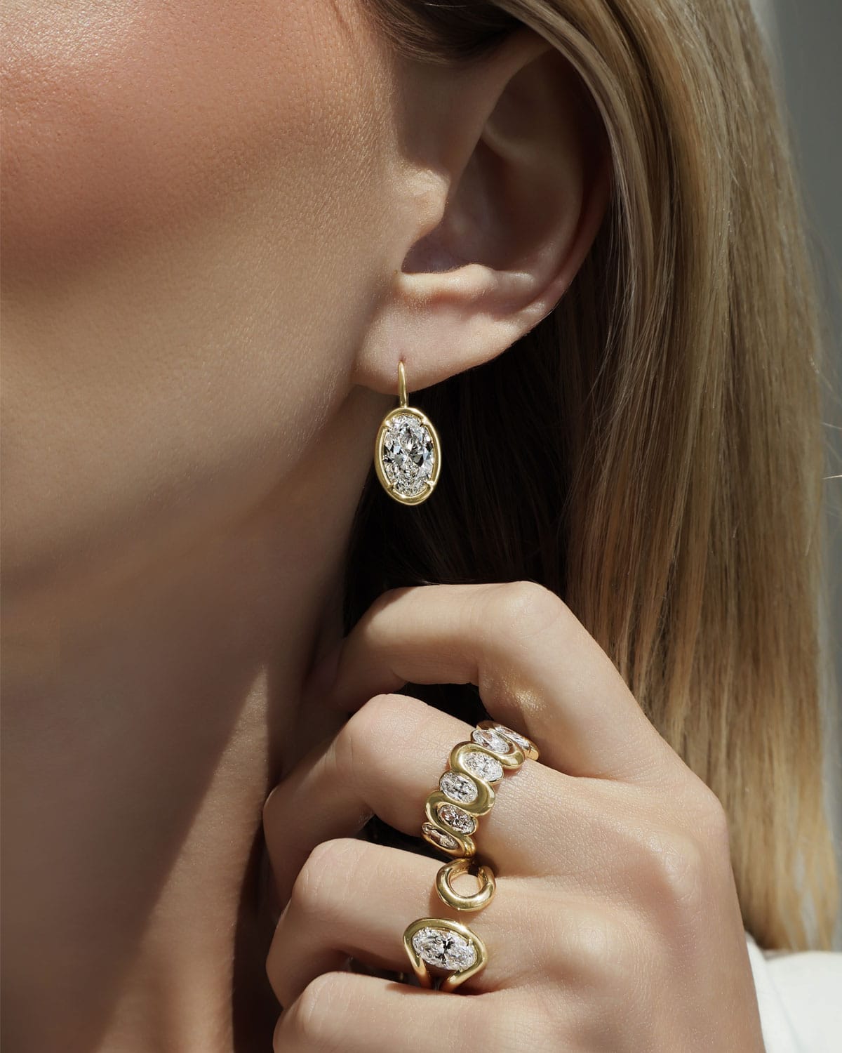 A close-up lifestyle shot of a model wearing a gold oval drop earring with a clear gemstone and matching gold rings on her fingers.