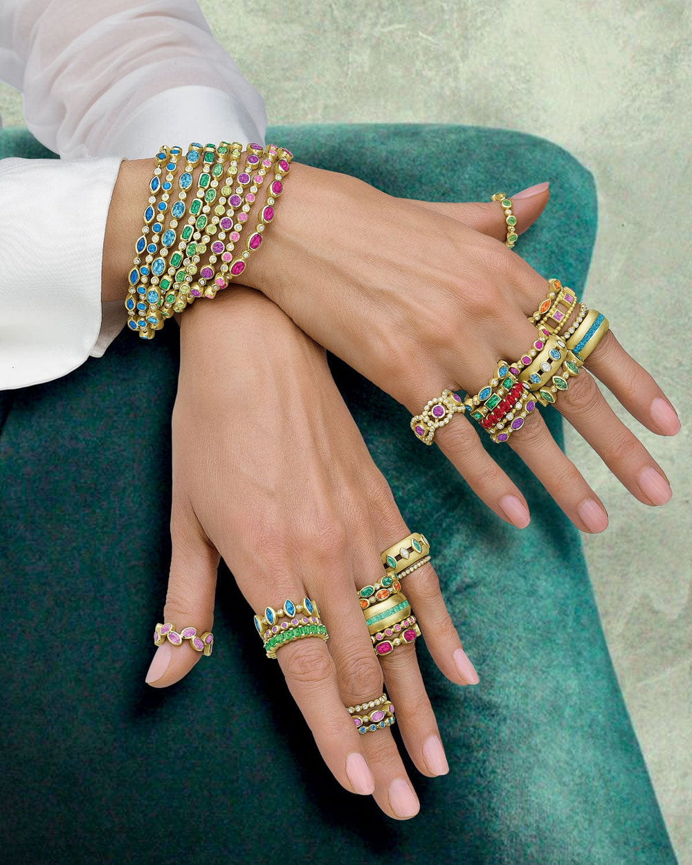 Close-up of hands wearing multiple colorful gemstone rings and bracelets on a green surface.