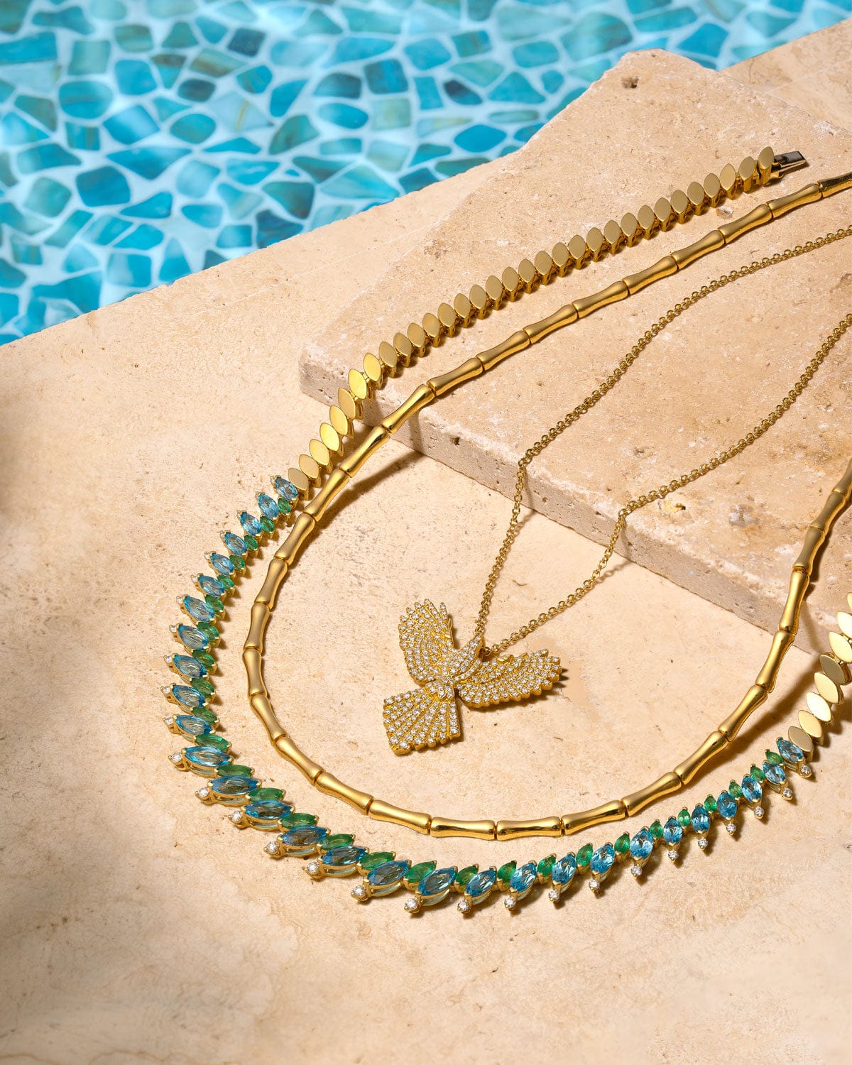 Gold necklace with green gemstones on a stone surface with a pool in the background