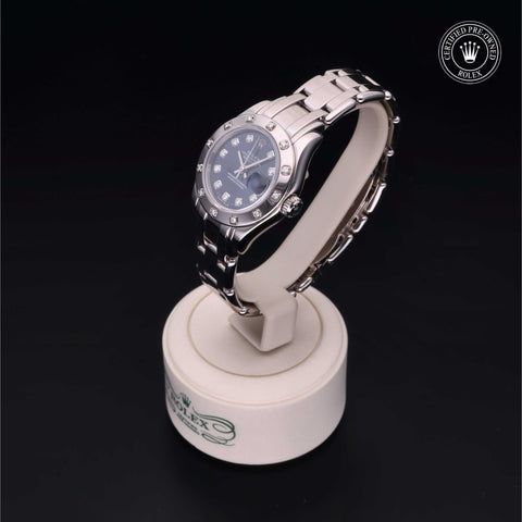 Silver wristwatch with a blue dial set with diamonds, displayed on a white stand against a black background. Rolex logo is visible in the upper right corner.