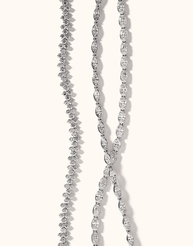 Two white gold-toned diamond tennis necklaces displayed vertically on a light cream background.