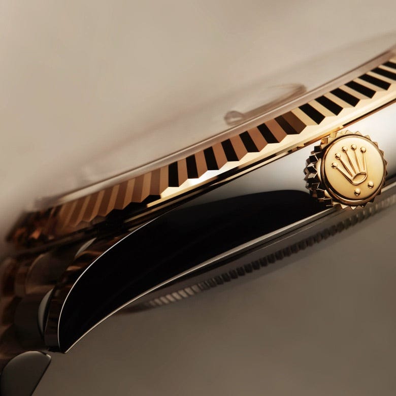 A close-up shot of a luxury wristwatch focusing on the gold crown and fluted bezel. The crown bears an embossed logo, and the bezel displays sharp, detailed ridges. The watch case has a polished metallic finish, reflecting light beautifully.