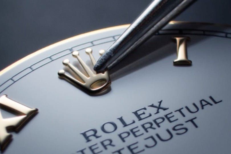 Close-up image of a Rolex watch face with a pen pointing towards the Rolex crown logo near the 12 o'clock mark. The watch's dial features the text 