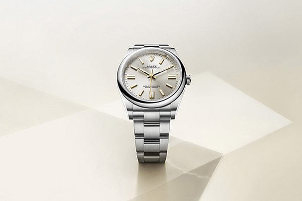 A sleek silver Rolex wristwatch is displayed against a minimalist geometric background. The watch features a stainless steel band and a round face with a light silver dial, gold hour markers, and hands. The Rolex logo is visible on the dial.