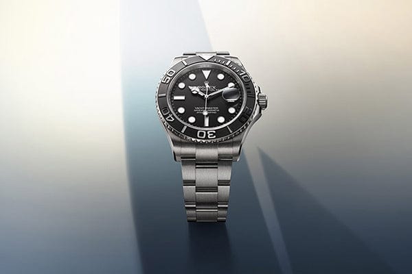 A luxury stainless steel wristwatch with a black dial and band, featuring luminous hour markers and hands. The watch is displayed upright on a gradient background that transitions from light to dark.