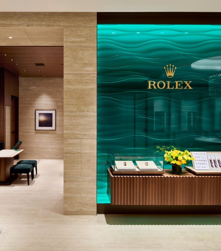 Luxury watch store interior featuring beige marble walls and flooring. Display cases with green backlighting hold watches, and a green panel with the Rolex logo dominates the scene. Dark green seating and a bouquet of yellow flowers add elegance.