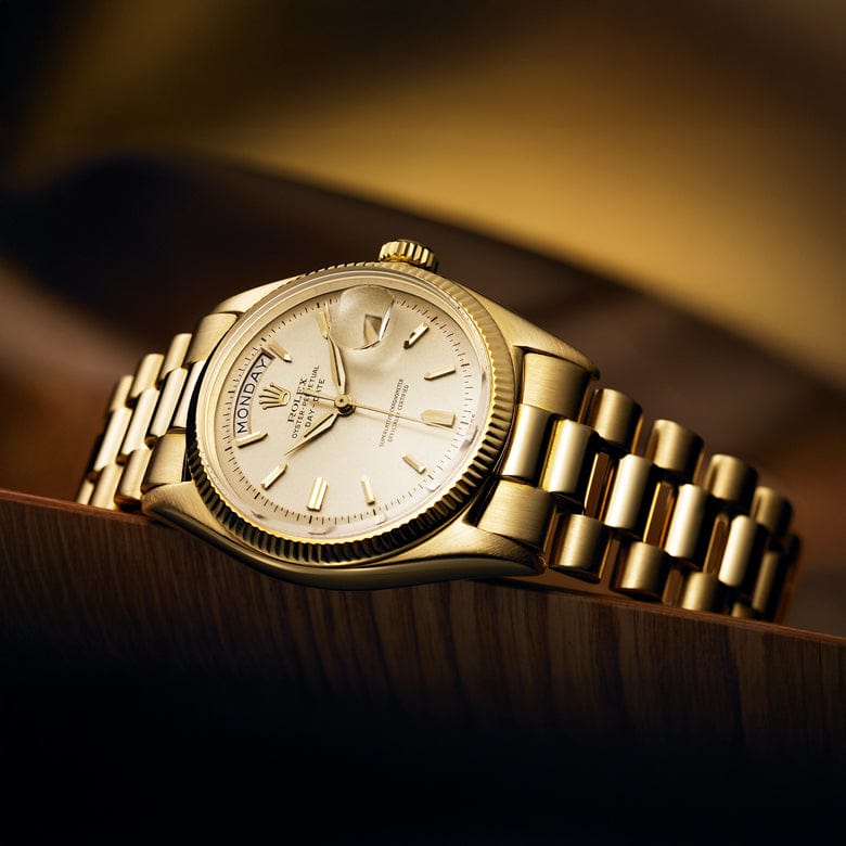 A gold wristwatch with a metallic bracelet, featuring a light-colored dial and gold hour markers, resting on a wooden surface against a warm, blurred background.