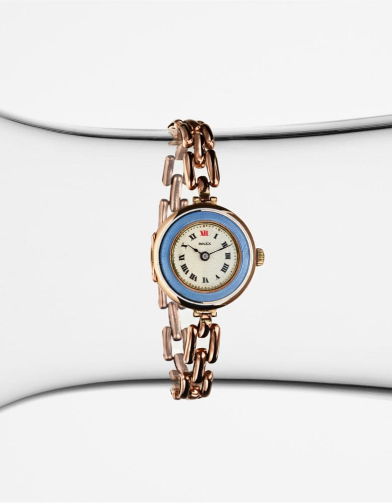 A vintage wristwatch with a round face, displaying roman numerals, and a blue bezel. The watch features a unique metallic link band, with alternating gold and silver tones. It is set against a clean, modern white and grey background.