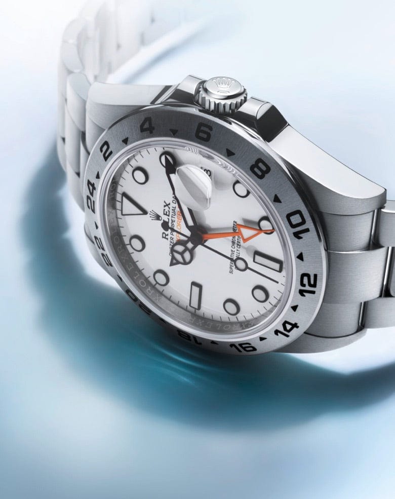 Close-up of a Rolex Explorer II watch with a stainless steel band. The watch features a white dial with black hour markers, a date window, and a bright orange 24-hour hand. The crown displays the Rolex logo, and the bezel has 24-hour markings.