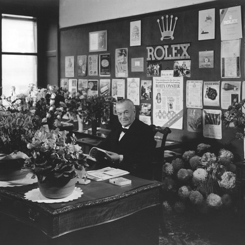 A black-and-white photo of an older man in a suit seated at a desk covered with large flower arrangements. The wall behind him displays various Rolex advertisements and a large Rolex sign with a crown above it. The man is holding a book and smiling.