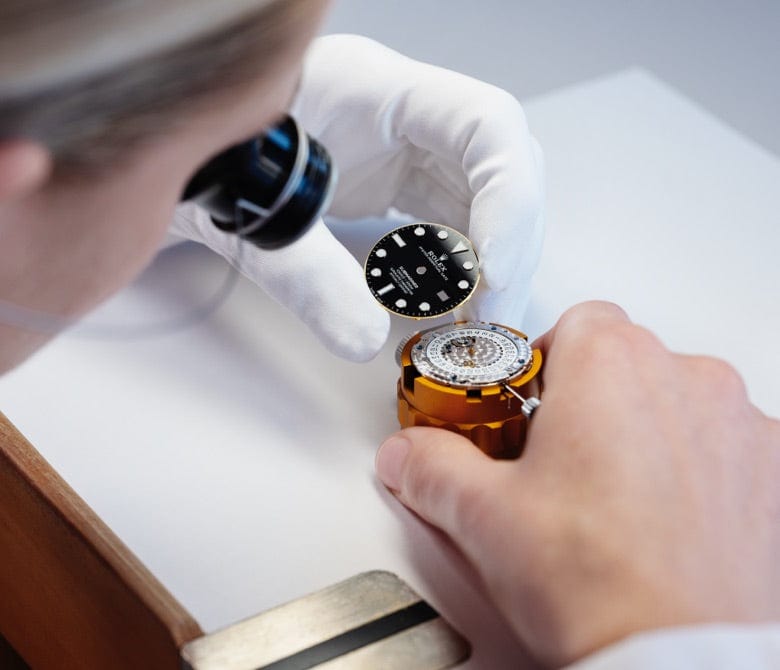 A close-up of a person wearing a white glove and using magnification tools while assembling a watch. They hold the black watch face with white details above the watch mechanism set in a golden holder, demonstrating precision craftsmanship.