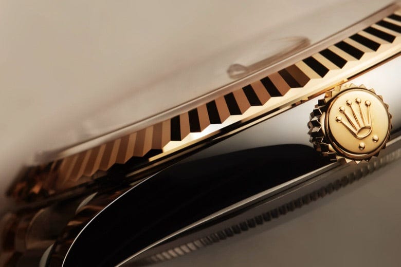 Close-up of a luxury watch showing intricate details of the gold crown, fluted bezel, and a polished side. The crown features a logo with a crown symbol, indicative of a prestigious brand. The image highlights the watch's craftsmanship and sophisticated design.