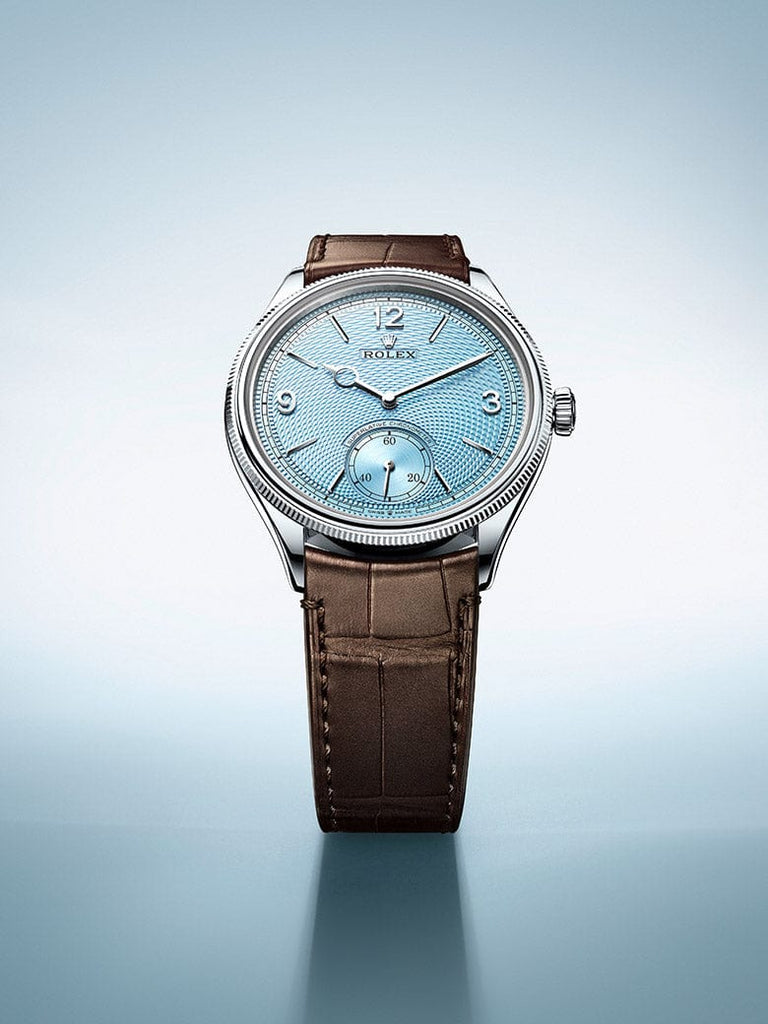 A luxury wristwatch with a light blue dial, metallic bezel, and brown leather strap is displayed against a gradient blue background. The watch face features Arabic numerals, a small second hand dial, and the brand name 