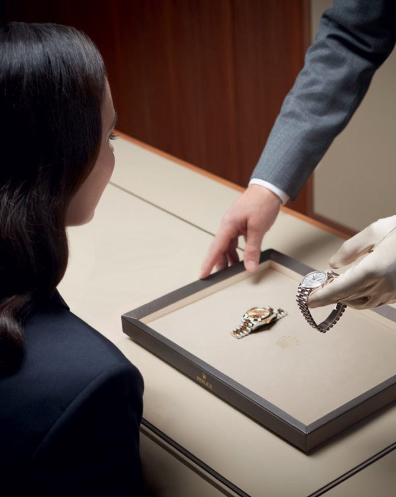 A person with long dark hair sits at a table while another person, wearing a grey suit and gloves, presents a luxury watch from a tray containing another watch. The scene is set in a high-end retail environment with a focus on elegant timepieces.
