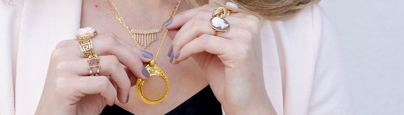 A woman wearing stunning vintage necklaces holds a gold chain with her hands laden with a selection of gold vintage rings