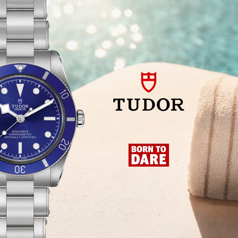 A Tudor watch with a blue dial and silver stainless steel bracelet, shown alongside the Tudor logo and a red 'Born to Dare' badge on a beach-themed background with sand and water.