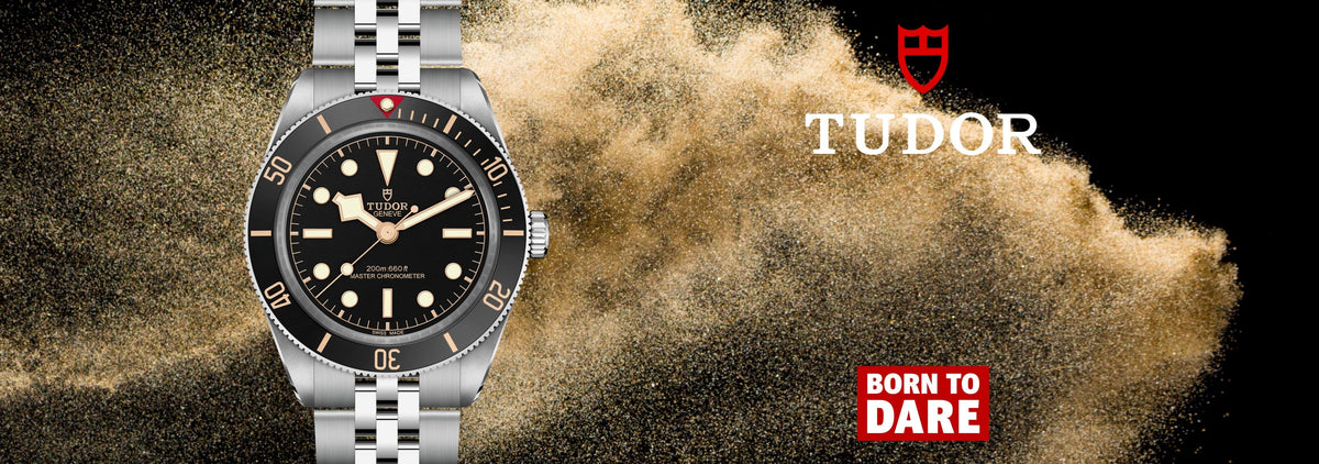 A Tudor watch with a black dial and silver-tone metal bracelet, shown against a dramatic background of sand explosion on black. The Tudor brand name and red shield logo are visible on the right, with a 'Born to Dare' badge at the bottom right.