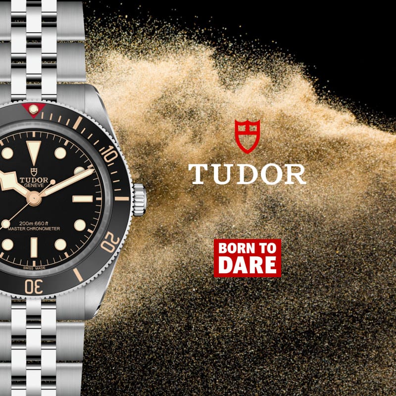 A Tudor wristwatch with a silver metal bracelet and black dial is shown alongside a dramatic burst of sand on a black background, with the Tudor logo and 'Born to Dare' text.