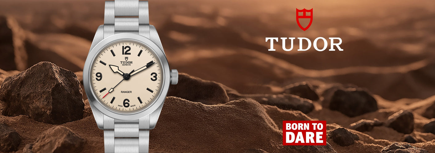 A silver-tone Tudor watch with a cream dial and black numerals, shown standing on rocky desert terrain with a warm brown background. The Tudor logo appears in the upper right, with a red shield icon and the slogan 'Born to Dare' in the lower right.