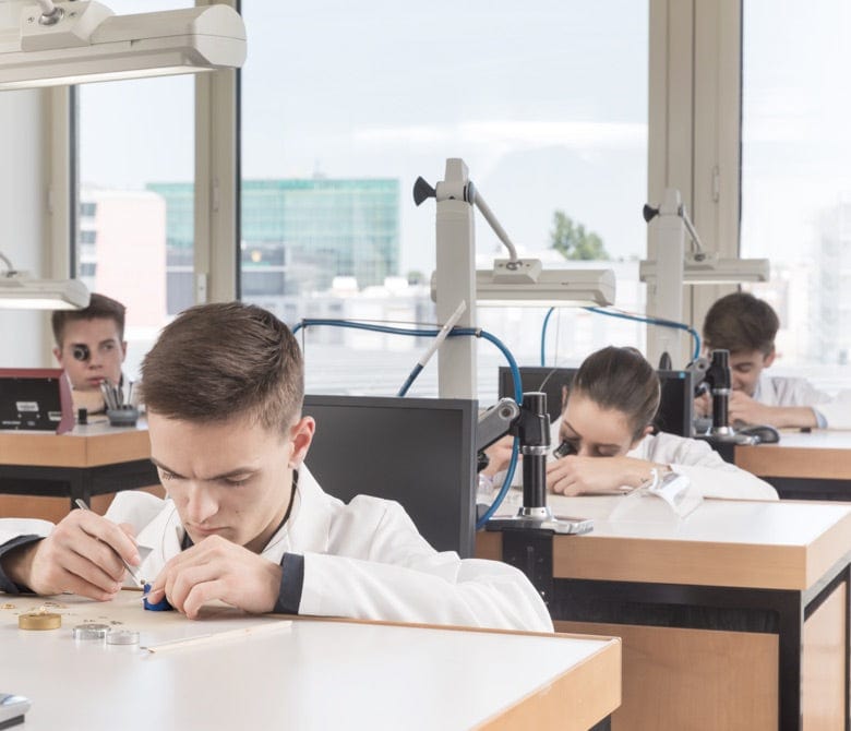 Students in white lab coats intensely focusing on small electronic components at a classroom workspace. The classroom is equipped with various tools and equipment.